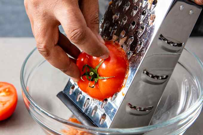 A hand pulps a tomato on the big holes of a box grater.